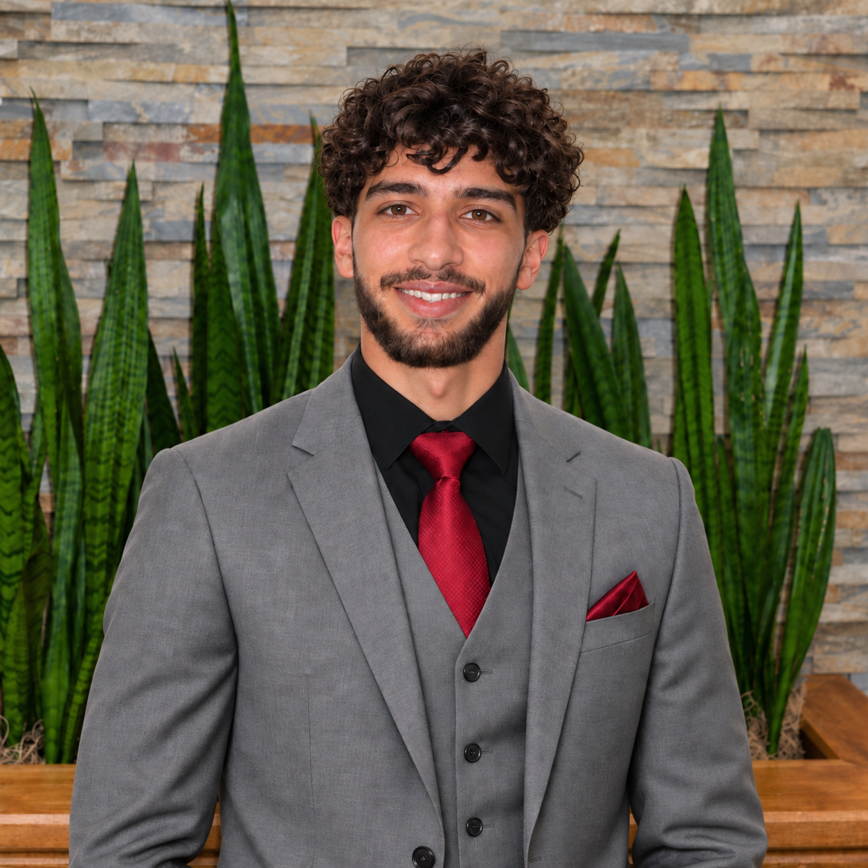 Matthew Rondon in a gray suit, standing in front of a stone wall and plants